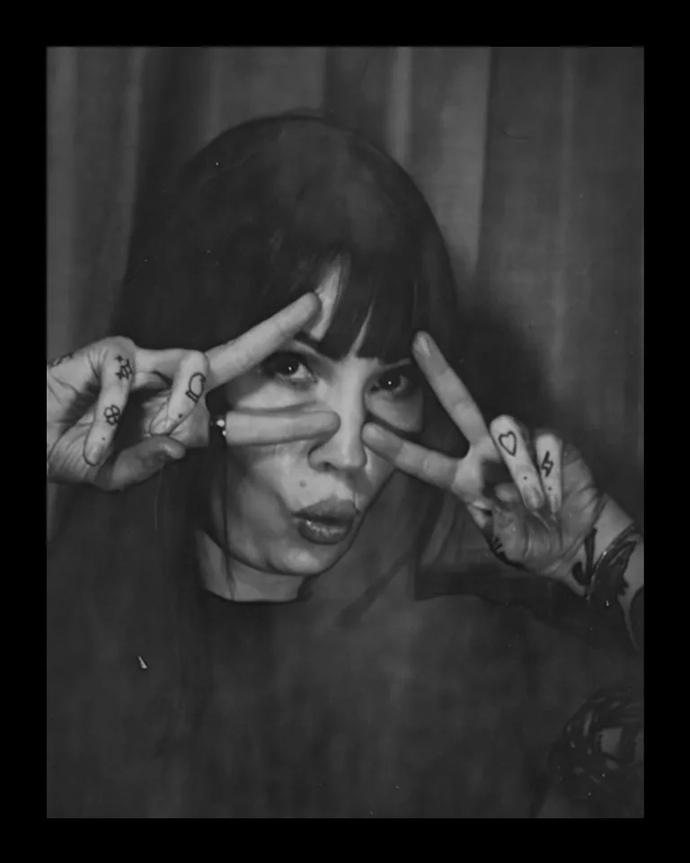 A black and white photo of a woman with dark hair and bangs, making a playful face with her lips puckered. She has tattoos on her hands and fingers, and is framing her eyes with her fingers in a peace sign gesture.