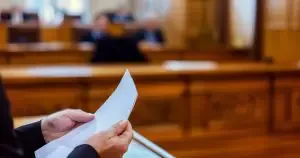 A person holding papers stands at a wooden podium in a courtroom.