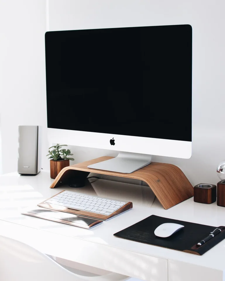 Modern workspace with an Apple iMac on a wooden stand, white keyboard, mouse, small potted plant, and decorative items on a white desk.